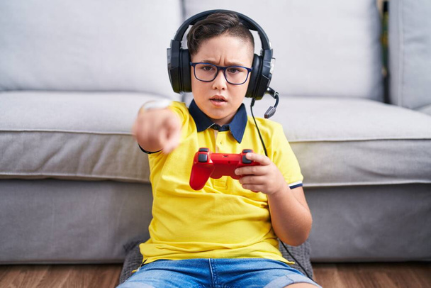 Junger hispanischer Junge, der Videospiel spielt und einen Controller mit Kopfhörern trägt, der unzufrieden und frustriert in die Kamera zeigt, wütend und wütend auf dich  - Foto, Bild