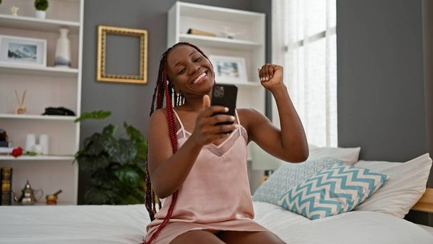 Afroamerykanka za pomocą smartfona siedzi na łóżku świętując w sypialni - Zdjęcie, obraz