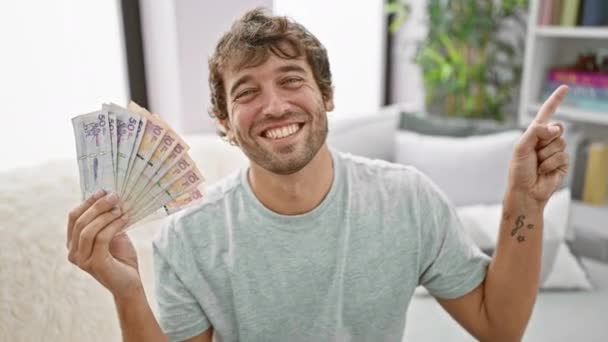 Χαρούμενος νεαρός άνδρας στο σπίτι, κρατώντας κολομβιανά πέσος, δείχνοντας με αυτοπεποίθηση το πλάι με ένα χαρούμενο χαμόγελο - Πλάνα, βίντεο