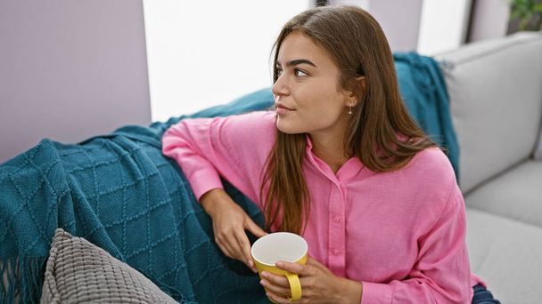Derin düşüncelere dalmış genç, güzel bir İspanyol kadın, evdeki kanepede rahatça oturuyor, sabah kahvesinin tadını çıkarıyor, rahatlamış konsantrasyonunu ve yaşam tarzını ifade ediyor. - Fotoğraf, Görsel