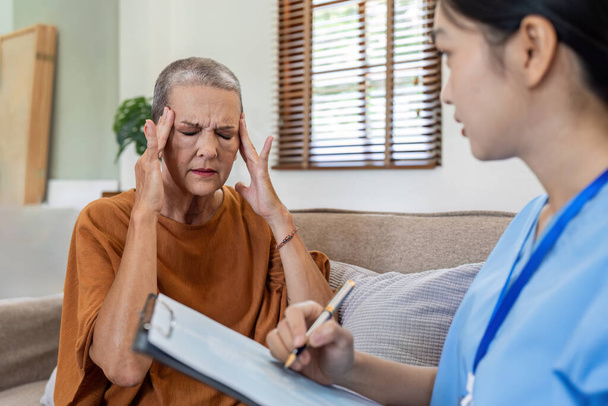 Kanepede başı ağrıyan yaşlı bir kadın ve ev sigortası olan bir hemşireyle konuşuyor ve koltukta baş ağrısı olan bir kadın.. - Fotoğraf, Görsel