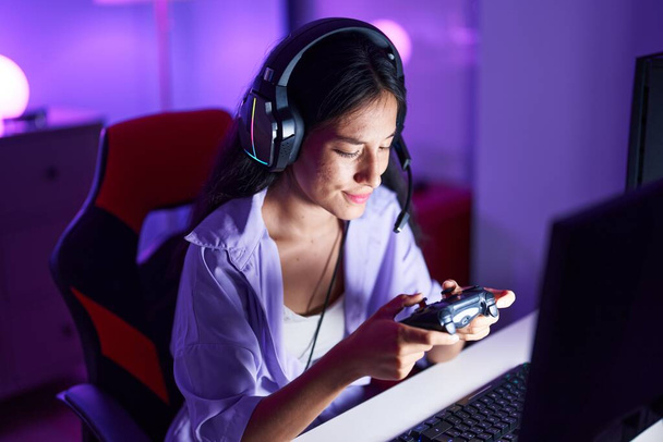 Young beautiful hispanic woman streamer playing video game using joystick at gaming room - Foto, imagen
