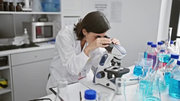 Jeune femme scientifique hispanique énergique avec une belle coiffure, absorbée dans sa recherche médicale, prenant méticuleusement des notes tout en regardant à travers un microscope dans un laboratoire animé. - Séquence, vidéo