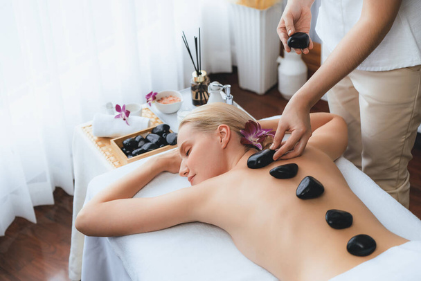 Hot stone massage at spa salon in luxury resort with day light serenity ambient, blissful woman customer enjoying spa basalt stone massage glide over body with soothing warmth. Quiescent - Photo, Image