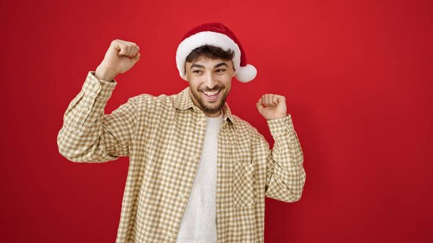 Hombre árabe joven con sombrero de Navidad con expresión alegre sobre fondo rojo aislado - Foto, Imagen