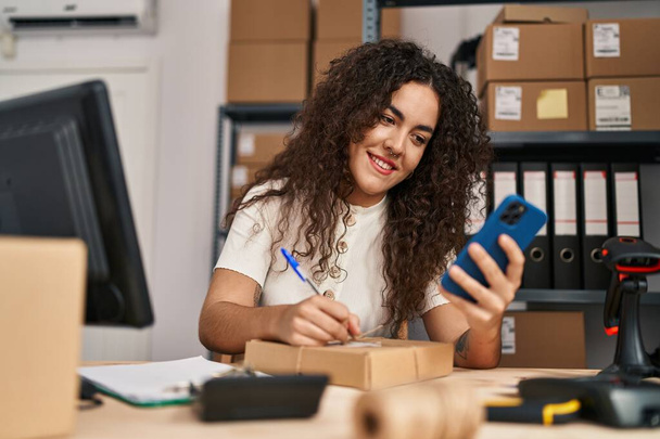 Fiatal gyönyörű spanyol nő e-kereskedelmi üzletember segítségével smartphone írás csomag az irodában - Fotó, kép