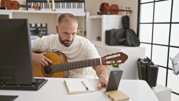 Jóképű fiatal kaukázusi férfi zenész lekötött online klasszikus gitár lecke zenei stúdióban, kecsesen játszik spanyol akusztikus hangszer, szeme az okostelefon képernyőjén - Felvétel, videó