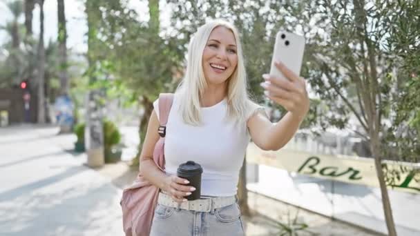 Vrolijke jonge blonde vrouw vol vertrouwen genieten van haar hete koffie terwijl ze glimlacht helder in het park, absorberen van het zonlicht van de stad, allemaal verpakt in een boeiende online video gesprek op haar telefoon. - Video