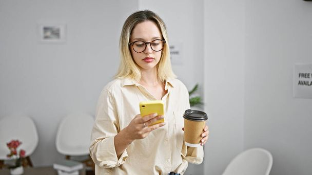 Jonge blonde vrouw zakenman met behulp van smartphone het drinken van koffie op kantoor - Foto, afbeelding