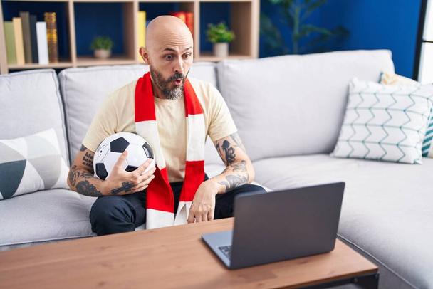 Іспаномовний чоловік з татуюваннями дивиться футбольний матч хуліган тримає м'яч на ноутбуці наляканий і вражений відкритим ротом для несподіванки, обличчя віри  - Фото, зображення