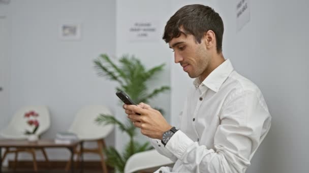 Young hispanic man using smartphone sitting on chair at waiting room - Materiał filmowy, wideo