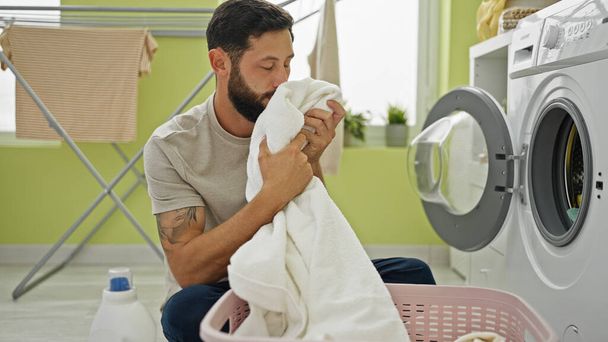 Jonge Latijns-Amerikaanse man wassen van kleding ruiken schone handdoek in wasruimte - Foto, afbeelding