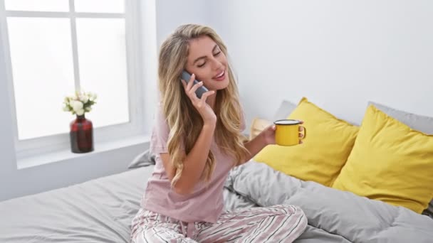 Mutlu genç bir kadın, yatakta akıllı telefonuyla rahatça konuşurken kahvesini yudumluyor. Sabahları rahat yatak odasında dinleniyor. - Video, Çekim