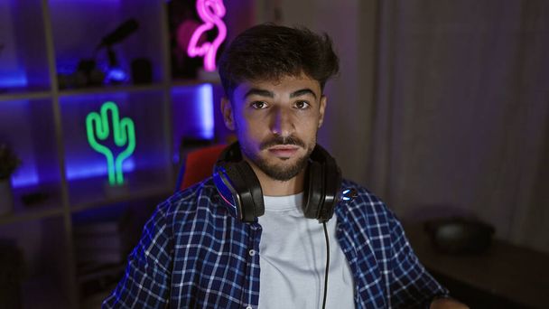 Konzentriert im Spiel, junger arabischer Mann, ein ernst dreinblickender Streamer, der Kopfhörer in einem schummrigen Spielzimmer trägt, vertieft in eine futuristische virtuelle Spielwelt - Foto, Bild
