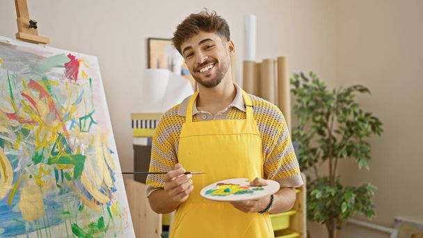 Gülümseyen Arap genç adam resim atölyesinde boya fırçası yeteneğini sergiliyor, resim hobisini ve eğitimini özgüvenle kucaklıyor. - Fotoğraf, Görsel