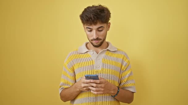 Cheerful young arab man exuding confidence, celebrating a win on his smartphone, smiling broadly against an isolated yellow background - Footage, Video