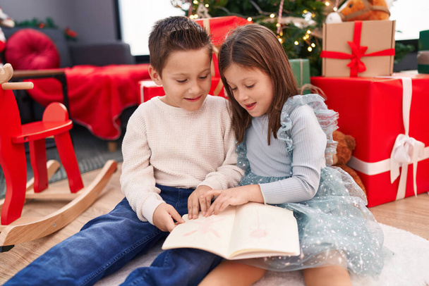 Очаровательный мальчик и девочка читают книгу, празднуя Рождество дома - Фото, изображение