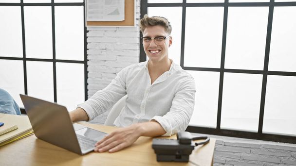 Счастливый и уверенный в себе, молодой кавказский человек, работающий над своим ноутбуком на рабочем месте в помещении. Деловой работник с непоколебимой уверенностью и сияющей улыбкой. - Фото, изображение