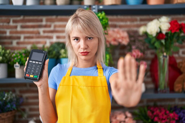 Mulher caucasiana jovem trabalhando na florista loja segurando dataphone com a mão aberta fazendo sinal de parada com expressão séria e confiante, gesto de defesa  - Foto, Imagem