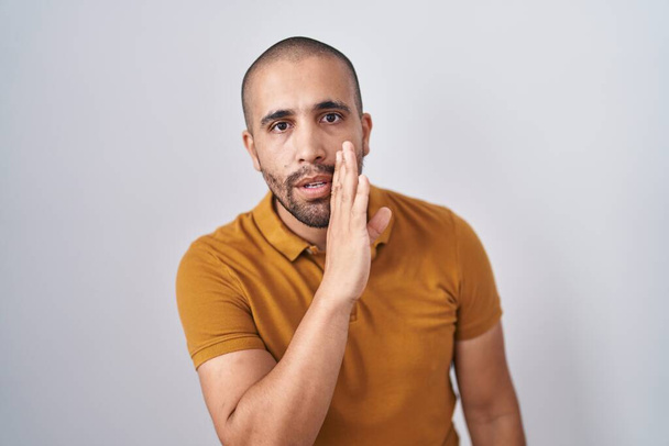 Hispanic man with beard standing over white background hand on mouth telling secret rumor, whispering malicious talk conversation  - Фото, изображение