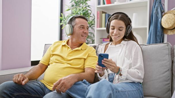W domu kochający ojciec i córka przytulnie siedzą na kanapie, słuchając ulubionej muzyki, uśmiechając się rozświetlając pokój, delektując się więzią, techno-gadżet audio lejąc zabawy i szczęścia. - Zdjęcie, obraz