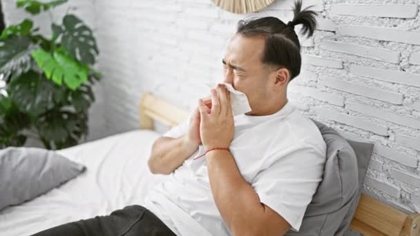 Beteg, fiatal kínai férfi, aki kovid-19-es fertőzésben szenved, az ágyon ül, a hálószobájában tüsszent, az influenza ellen az egészségügyi ellátás támogatásával küzd. - Felvétel, videó