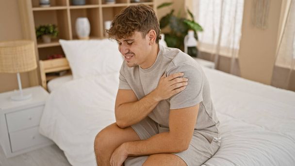 Worried young hispanic man, in pyjamas, suffering intense shoulder pain. unhappy, he lies awake in his home, touching his injured arm on his bed in the morning. - Photo, Image