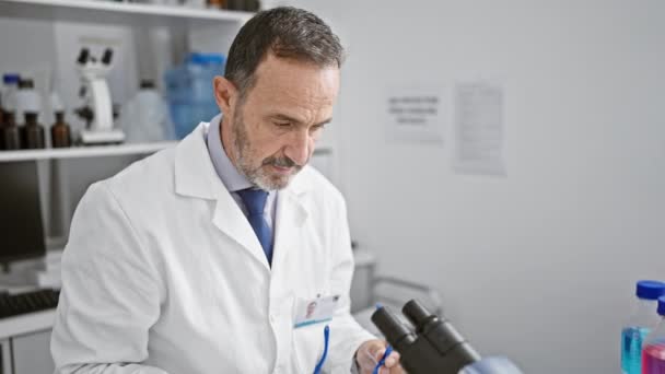 Homme d'âge moyen avec des cheveux gris scientifique portant des lunettes de sécurité en utilisant un microscope au laboratoire - Séquence, vidéo