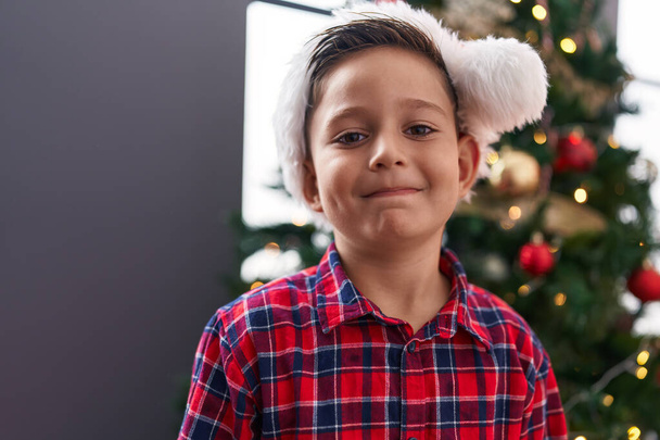 Αξιολάτρευτο ισπανόφωνο αγόρι χαμογελά σίγουρος στέκεται δίπλα στο χριστουγεννιάτικο δέντρο στο σπίτι - Φωτογραφία, εικόνα