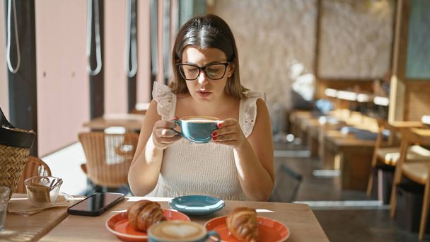Gyönyörű spanyol nő élvezi forró kávé ital belsejében napfényes kávézó, pihentető reggelinél, miközben szemüveget visel, ül lazán cafeteria asztal élvezi a reggeli nap. - Fotó, kép