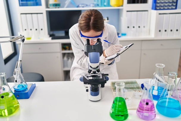 Jeune femme blonde scientifique utilisant le microscope écrit sur le document au laboratoire - Photo, image