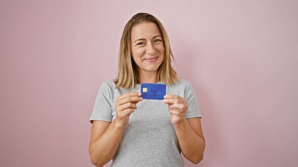 Fröhliche junge blonde Frau hält selbstbewusst ihre Kreditkarte in der Hand und strahlt Freude und finanzielle Zuversicht vor isoliertem rosa Hintergrund aus - Foto, Bild