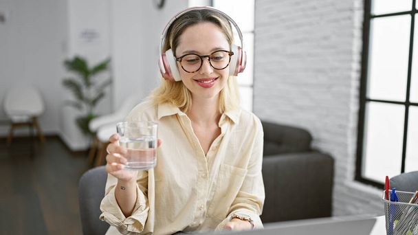Nuori blondi nainen liiketoiminnan työntekijä käyttää kannettavaa tietokonetta ja kuulokkeet juomavettä toimistossa - Valokuva, kuva