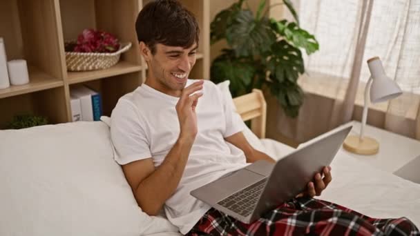 Joyeux jeune homme hispanique saluant lors d'une conversation d'appel vidéo en ligne heureux, assis sur le lit avec son ordinateur portable dans l'intérieur confortable de sa chambre à coucher à la maison - Séquence, vidéo
