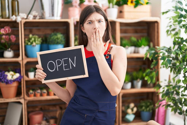 Young caucasian woman working at florist holding open sign covering mouth with hand, shocked and afraid for mistake. surprised expression  - Photo, Image