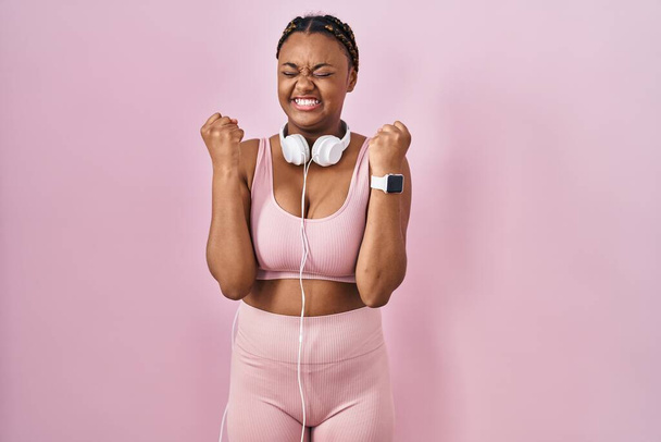 African american woman with braids wearing sportswear and headphones excited for success with arms raised and eyes closed celebrating victory smiling. winner concept.  - Photo, Image