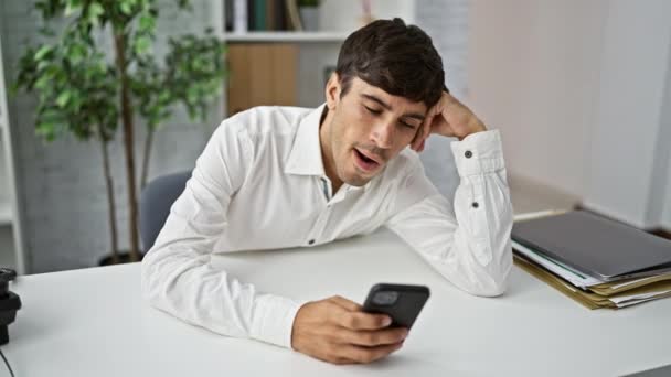 Εξαντλημένος νεαρός Ισπανός επιχειρηματίας αγωνίζεται να συγκεντρωθεί, χασμουριέται κοιτάζοντας smartphone στο γραφείο - Πλάνα, βίντεο