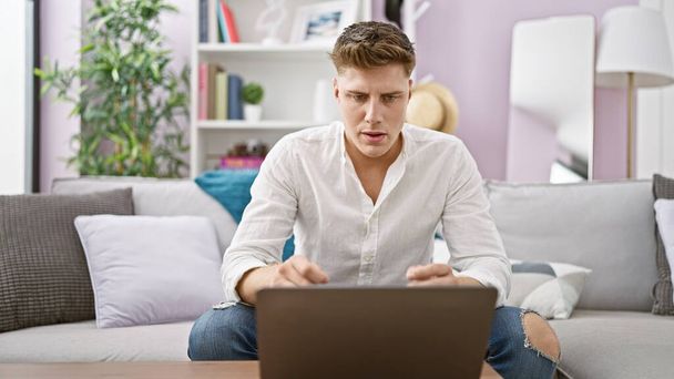 Орієнтований молодий кавказький чоловік сидить в приміщенні на дивані, використовуючи ноутбук, заглиблений в онлайн-технології вдома! красивий чоловік, абсолютно серйозно, серед інтер'єру своєї квартири. - Фото, зображення