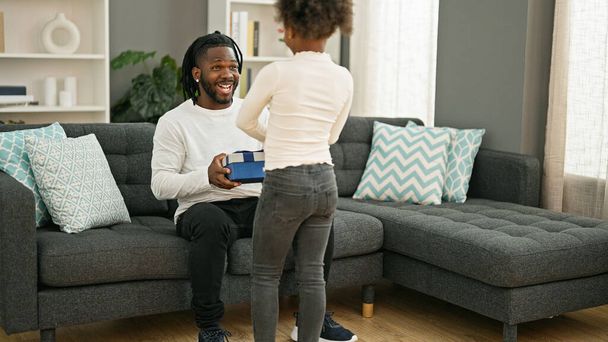 Afro-Amerikaanse vader en dochter verrassing met cadeau zitten op de bank glimlachen thuis - Foto, afbeelding