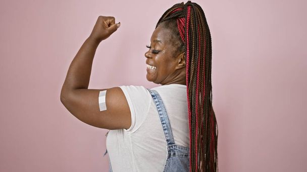 Zelfverzekerde Afro-Amerikaanse vrouw, sportvlechten en een pleister op haar arm, die vreugde uitstraalt met een sterk gebaar. haar mooie, vrolijke glimlach bovenop een geïsoleerde roze achtergrond, stralende positiviteit - Foto, afbeelding
