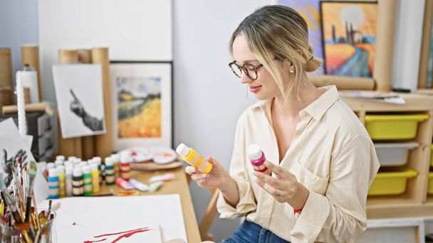 Junge blonde Künstlerin sitzt am Tisch und wählt Farbe lächelnd im Kunstatelier - Foto, Bild