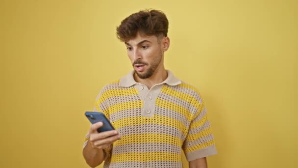 Jonge Arabier man, baard en al, verbaasd en breed-eyed, staan in verrassing als hij kijkt naar zijn smartphone, sms 'en weg op een geïsoleerde gele muur achtergrond. Wow, is dat een ongelooflijke boodschap! - Video
