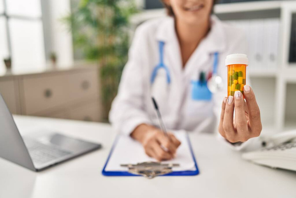 Mladá krásná hispánská žena lékař drží pilulky láhev psaní na dokument na klinice - Fotografie, Obrázek
