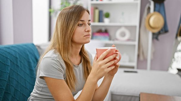 Ελκυστική νεαρή ξανθιά γυναίκα που κάθεται άνετα σε εσωτερικούς χώρους στο σαλόνι του διαμερίσματός της, εμπλουτίζοντας το πρωινό της χαλαρώνοντας και πίνοντας ένα ζεστό καφέ espresso ενώ αμφιβάλω. - Φωτογραφία, εικόνα