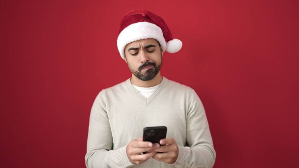 Junger hispanischer Mann mit Weihnachtsmütze und Smartphone mit unglücklichem Gesichtsausdruck vor isoliertem roten Hintergrund - Foto, Bild