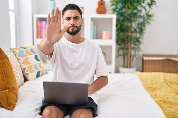 Joven hispano usando computadora portátil sentado en la cama con la mano abierta haciendo stop sign con expresión seria y segura, gesto de defensa  - Foto, imagen