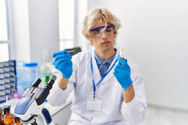 Joven científico hombre rubio sosteniendo botella y muestra con pinza en el laboratorio - Foto, Imagen
