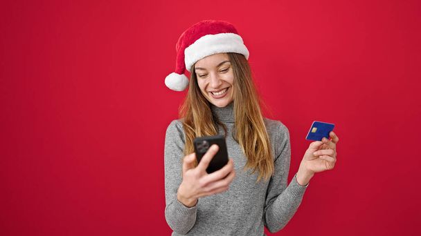 Mujer caucásica joven haciendo compras en línea de Navidad con teléfono inteligente sobre fondo rojo aislado - Foto, imagen