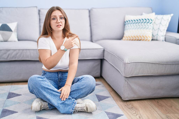 Nuori valkoihoinen nainen istuu lattialla olohuoneen osoittaa syrjään huolissaan ja hermostunut etusormi, huolissaan ja yllättynyt ilme  - Valokuva, kuva