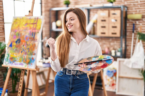 Fiatal kaukázusi nő a művészeti stúdióban palettát tartva, hüvelykujjal felfelé mosolyogva, boldogan nyitott szájjal.  - Fotó, kép